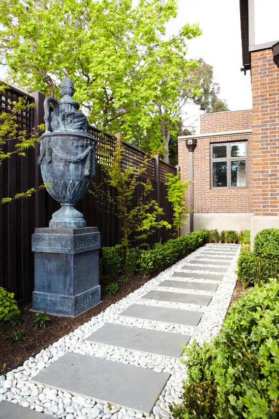 Bluestone stepping pavers edged with white pebbles, leading past a lead urn on a plinth beside a brick house